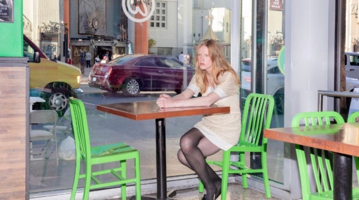 A woman with blonde hair wearing a brown dress and black tights and shoes sits at a table a table alone by a window